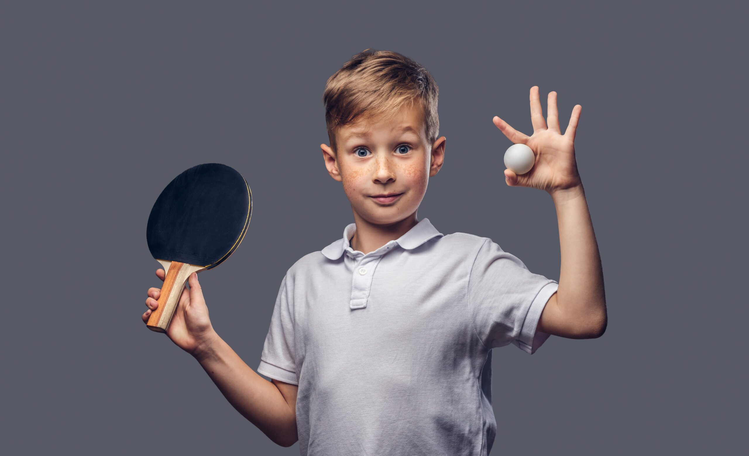 Cute redhead boy dressed in a white t-shirt holds a ping-pong racquet and ball in a studio